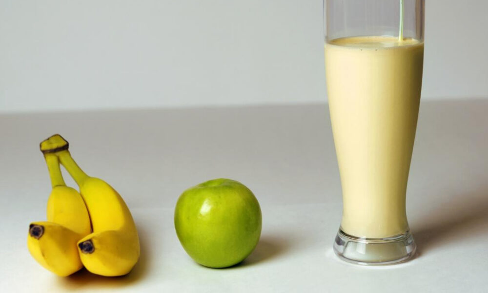 Bananas, a green apple, and a tall glass of creamy banana smoothie on a white surface.