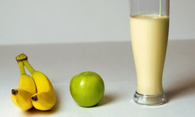 Bananas, a green apple, and a tall glass of creamy banana smoothie on a white surface.