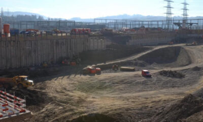 Large construction site with a deep excavated pit, dirt roads, and heavy machinery (dump trucks, excavator, and orange barriers) at the edge of the hole; distant industrial structures and power lines loom over the scene.