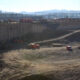 Large construction site with a deep excavated pit, dirt roads, and heavy machinery (dump trucks, excavator, and orange barriers) at the edge of the hole; distant industrial structures and power lines loom over the scene.