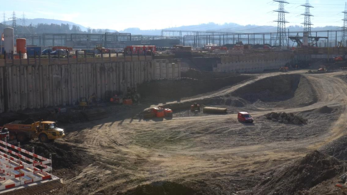 Large construction site with a deep excavated pit, dirt roads, and heavy machinery (dump trucks, excavator, and orange barriers) at the edge of the hole; distant industrial structures and power lines loom over the scene.