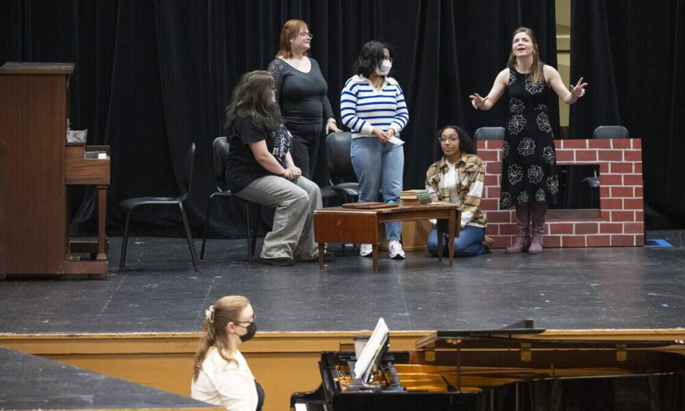Group of diverse women on a theater stage, one woman in a black dress speaks while others listen, piano in foreground.
