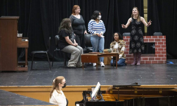 Group of diverse women on a theater stage, one woman in a black dress speaks while others listen, piano in foreground.