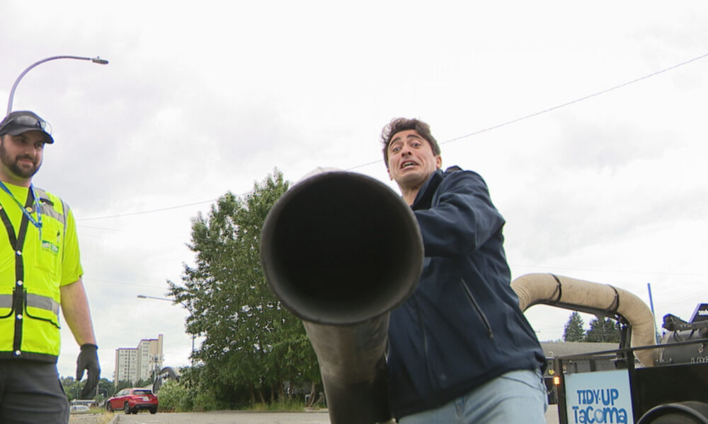 Two workers outdoors; one in a bright yellow safety vest watches as the other holds a large black pipe toward the camera on a street scene.
