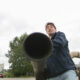 Two workers outdoors; one in a bright yellow safety vest watches as the other holds a large black pipe toward the camera on a street scene.
