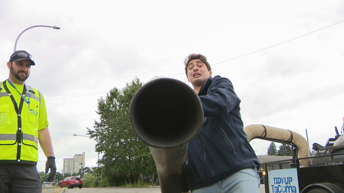 Two workers outdoors; one in a bright yellow safety vest watches as the other holds a large black pipe toward the camera on a street scene.