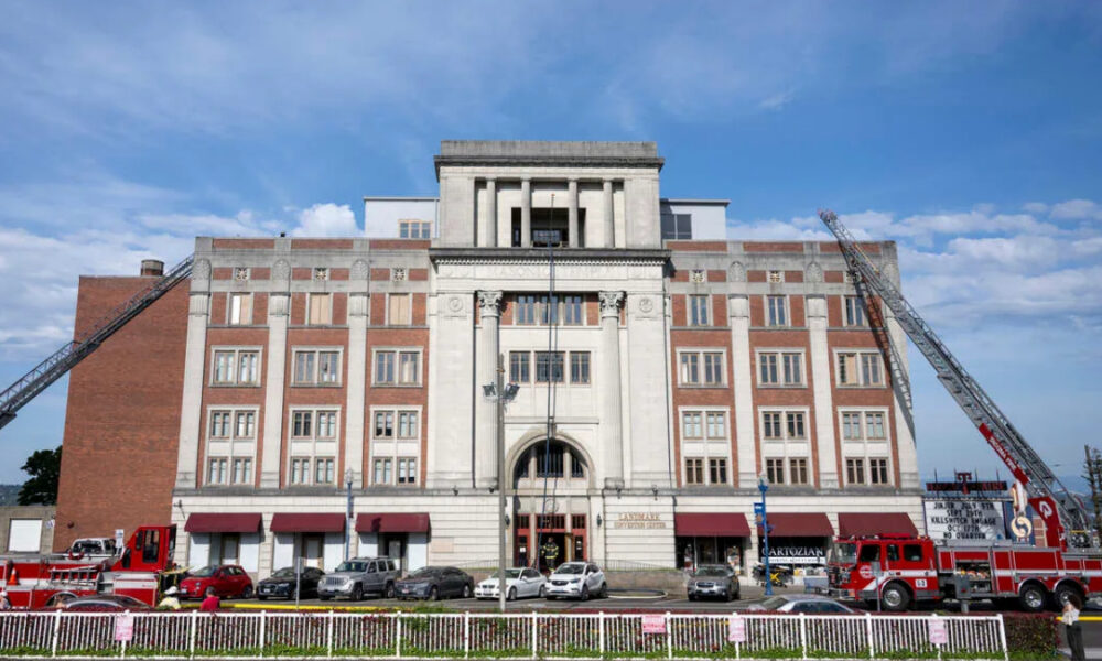 Fire department building with two extendable ladders leaning against the façade and red fire trucks parked in front on a sunny day.