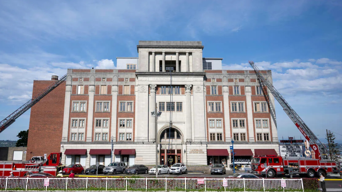 Fire department building with two extendable ladders leaning against the façade and red fire trucks parked in front on a sunny day.