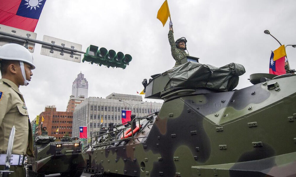 Soldier in a helmet raises a yellow flag from an open-turret on a camouflaged military tank during a city parade, with Taiwanese flags in the background.