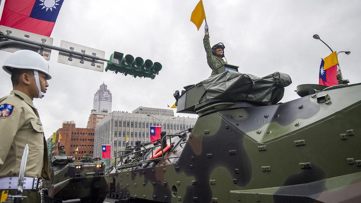 Soldier in a helmet raises a yellow flag from an open-turret on a camouflaged military tank during a city parade, with Taiwanese flags in the background.