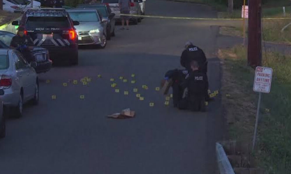 Two police officers kneel on a residential street surrounded by numerous yellow evidence markers and scattered items; police cars and caution tape in the scene.