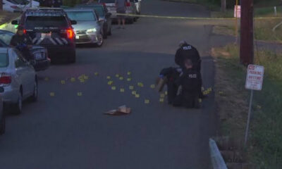Two police officers kneel on a residential street surrounded by numerous yellow evidence markers and scattered items; police cars and caution tape in the scene.