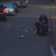 Two police officers kneel on a residential street surrounded by numerous yellow evidence markers and scattered items; police cars and caution tape in the scene.