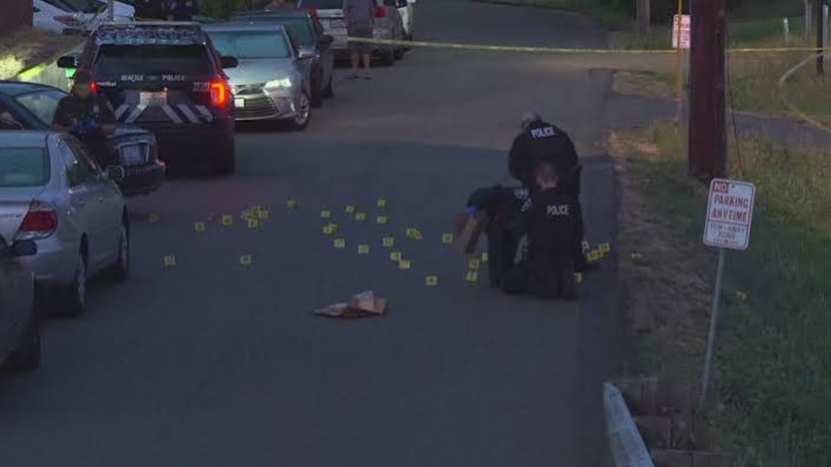 Two police officers kneel on a residential street surrounded by numerous yellow evidence markers and scattered items; police cars and caution tape in the scene.