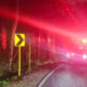 Night roadside scene with a yellow left-curve chevron sign beside a forest, and a fire truck with flashing red lights on a curved road central/right.
