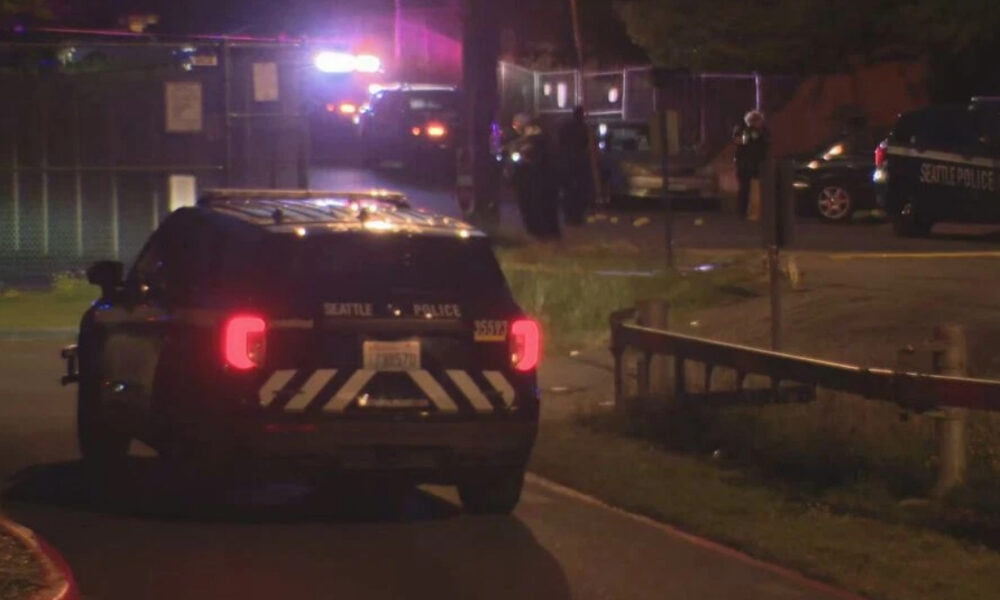 Nighttime scene with a Seattle Police SUV and flashing emergency lights parked on a street near a fenced area, with officers in the background.