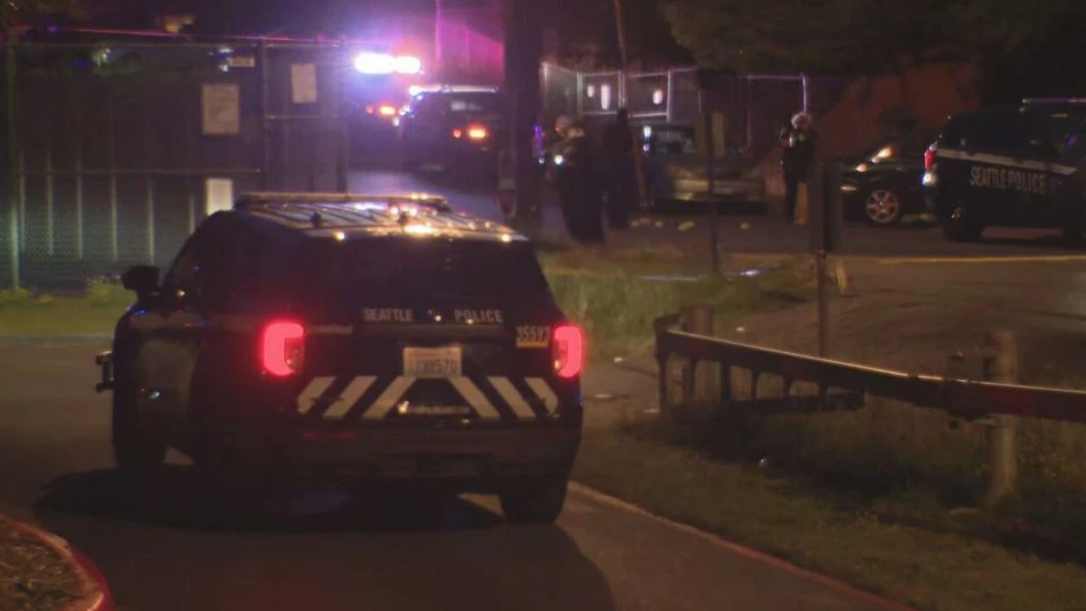 Nighttime scene with a Seattle Police SUV and flashing emergency lights parked on a street near a fenced area, with officers in the background.