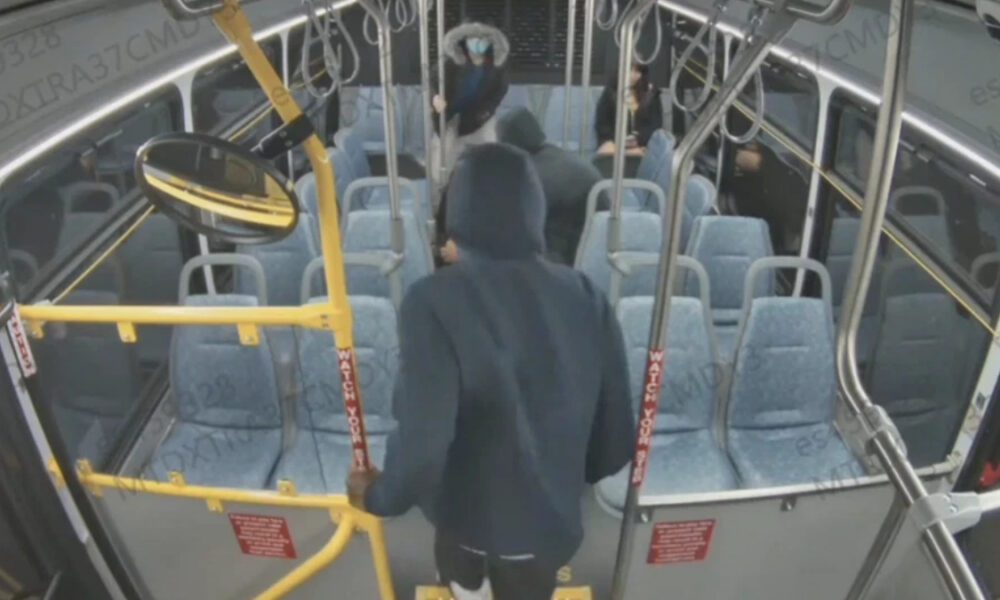Person in a dark hoodie boarding a city bus, seen from behind, with yellow gates and red 'Watch Your Step' straps near the front door.