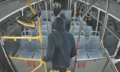 Person in a dark hoodie boarding a city bus, seen from behind, with yellow gates and red 'Watch Your Step' straps near the front door.