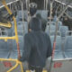 Person in a dark hoodie boarding a city bus, seen from behind, with yellow gates and red 'Watch Your Step' straps near the front door.