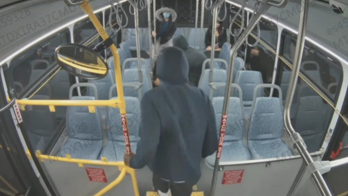 Person in a dark hoodie boarding a city bus, seen from behind, with yellow gates and red 'Watch Your Step' straps near the front door.