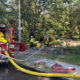 Fire engine with hoses connected to taps, firefighters in reflective gear near a tree-lined park area.