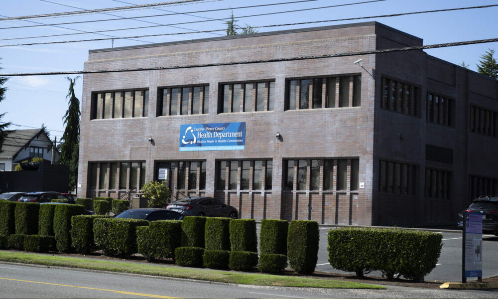 Brick two-story health department building with a blue sign, lined by trimmed hedges and parked cars in front.