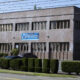 Brick two-story health department building with a blue sign, lined by trimmed hedges and parked cars in front.