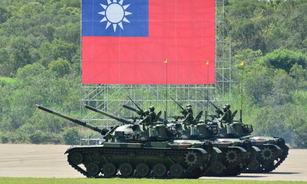 Armored tanks with soldiers atop, lined up on a parade ground under a giant Taiwan flag backdrop.