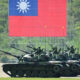 Armored tanks with soldiers atop, lined up on a parade ground under a giant Taiwan flag backdrop.