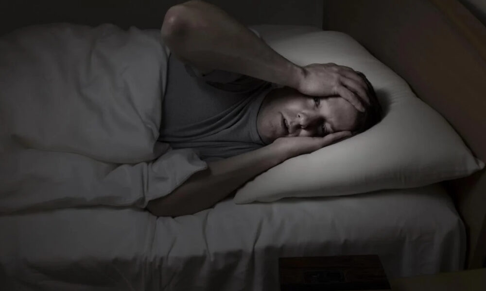 Man lying in bed at night, clutching his head with both hands in apparent headache or distress.