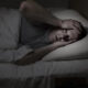 Man lying in bed at night, clutching his head with both hands in apparent headache or distress.