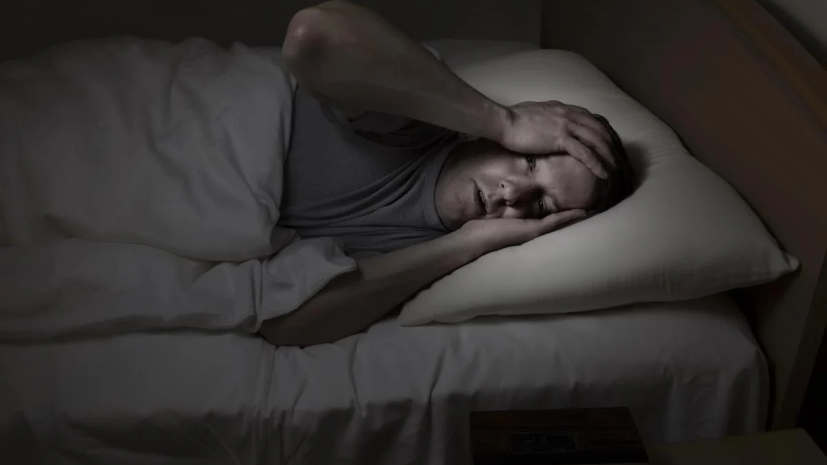 Man lying in bed at night, clutching his head with both hands in apparent headache or distress.