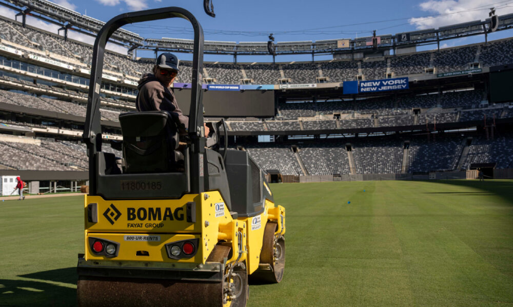 Yellow BOMAG roller with a worker driving it on a well-manicured stadium field, empty stands in the background and a blue sky above.
