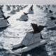 Row of identical white boat sculptures with black sails floating on the sea, arranged in a grid.
