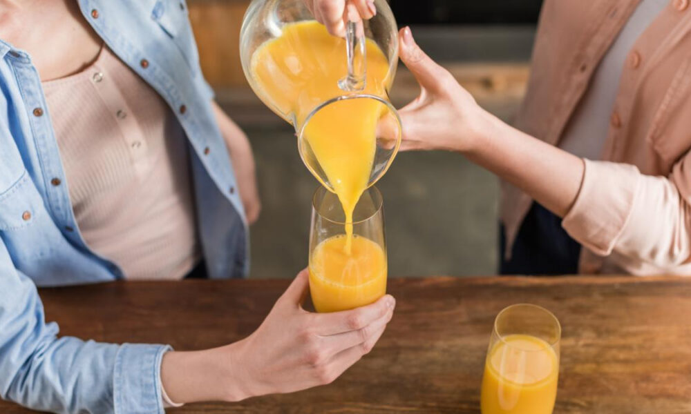 Two people pouring orange juice from a pitcher into a glass at a wooden table.