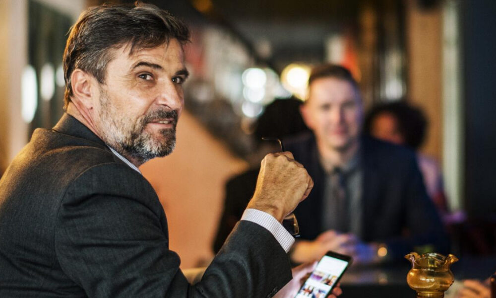 Middle-aged man in a dark blazer sits at a bar or restaurant, looking toward the camera while holding eyeglasses; a phone rests on the table in front, with blurred diners in the background.