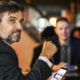 Middle-aged man in a dark blazer sits at a bar or restaurant, looking toward the camera while holding eyeglasses; a phone rests on the table in front, with blurred diners in the background.