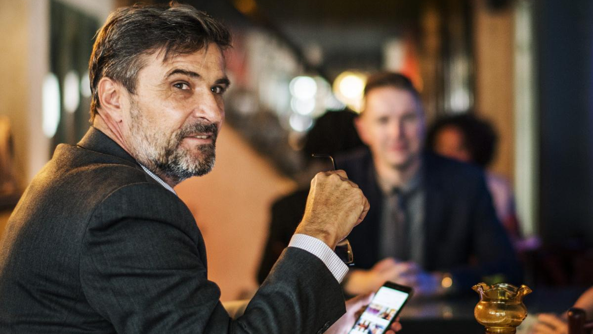 Middle-aged man in a dark blazer sits at a bar or restaurant, looking toward the camera while holding eyeglasses; a phone rests on the table in front, with blurred diners in the background.
