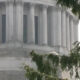 White marble neoclassical building with tall columns, partially obscured by green tree branches.