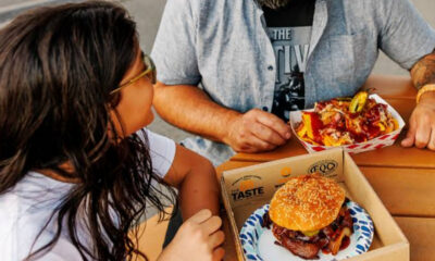 Two people sit at a wooden table sharing a meal: a sesame-seed hamburger in a cardboard box and fries on a plate.