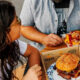 Two people sit at a wooden table sharing a meal: a sesame-seed hamburger in a cardboard box and fries on a plate.