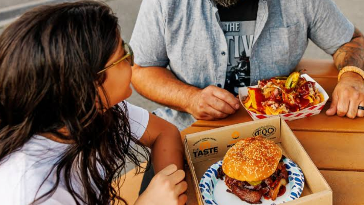Two people sit at a wooden table sharing a meal: a sesame-seed hamburger in a cardboard box and fries on a plate.
