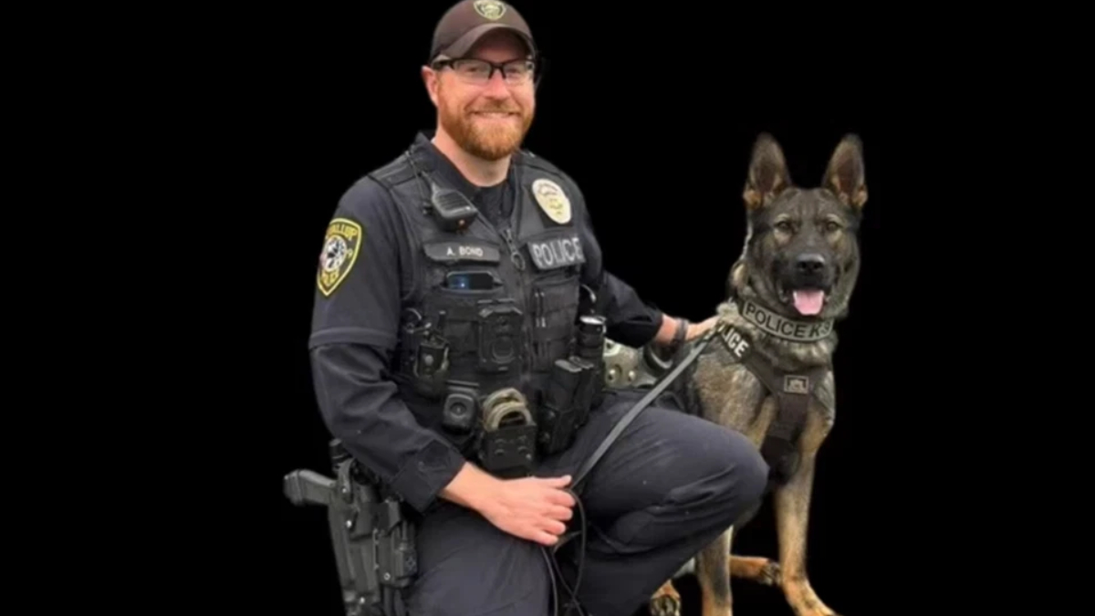 Police officer in tactical gear kneels beside a German Shepherd police dog against a black background.