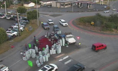 Aerial view of a roundabout with a white truck and many stacked white barrels, a red liquid spill on the pavement, and workers in safety vests nearby.