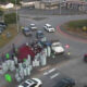 Aerial view of a roundabout with a white truck and many stacked white barrels, a red liquid spill on the pavement, and workers in safety vests nearby.