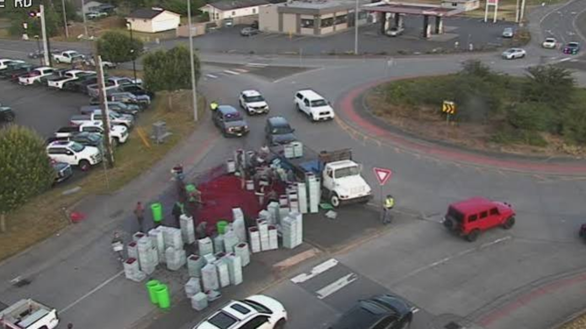Aerial view of a roundabout with a white truck and many stacked white barrels, a red liquid spill on the pavement, and workers in safety vests nearby.