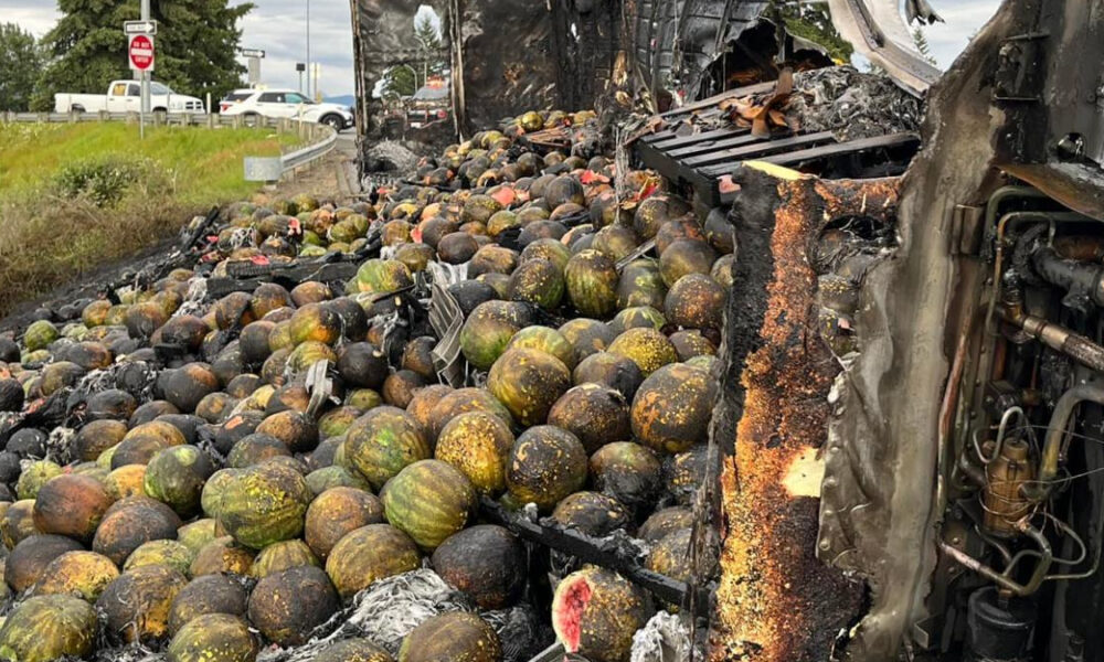 Watermelons and melons spilled across a highway after a crash, with a severely damaged truck on the right and emergency vehicles in the distance
