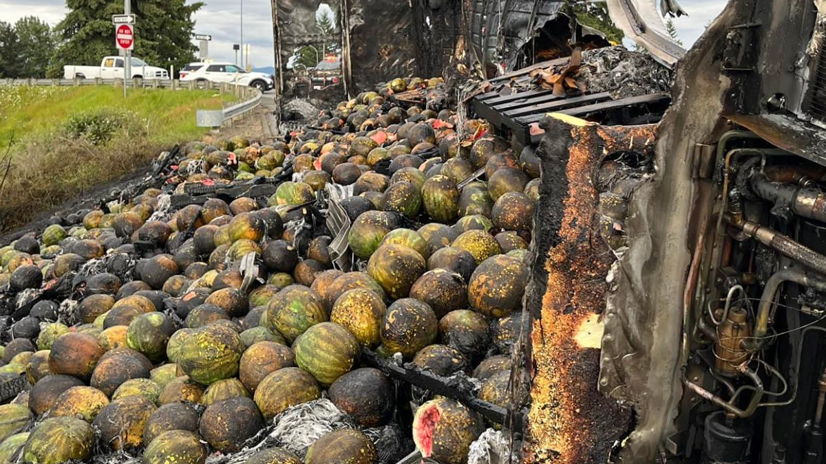 Watermelons and melons spilled across a highway after a crash, with a severely damaged truck on the right and emergency vehicles in the distance