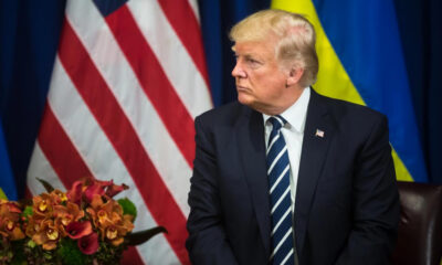 A man in a dark suit and striped tie sits at a desk with a flower arrangement, with American and blue-yellow flags in the background.